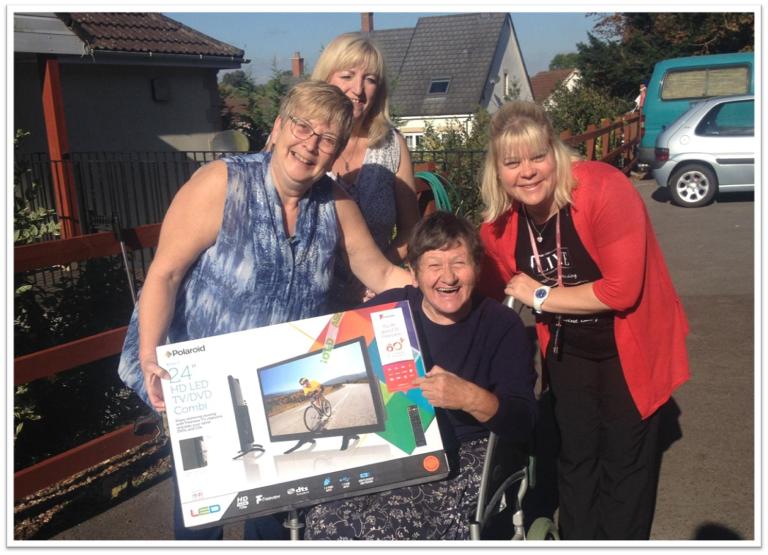 a lady sat down on a wheelchair surrounded buy three other ladies all happy holding a new TV box