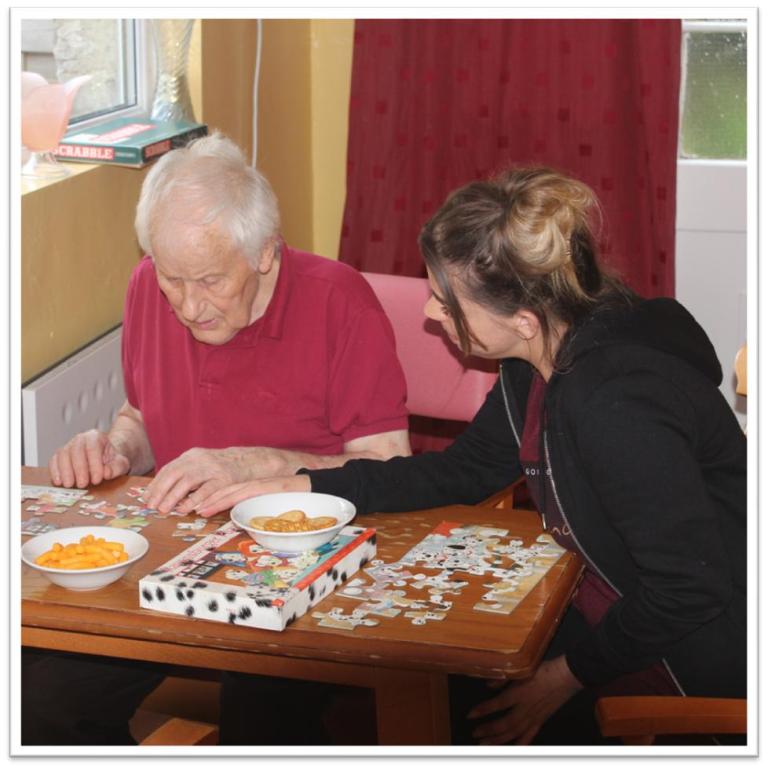 a woman helping an older man with a jigsaw puzzle whilst they eat carrots