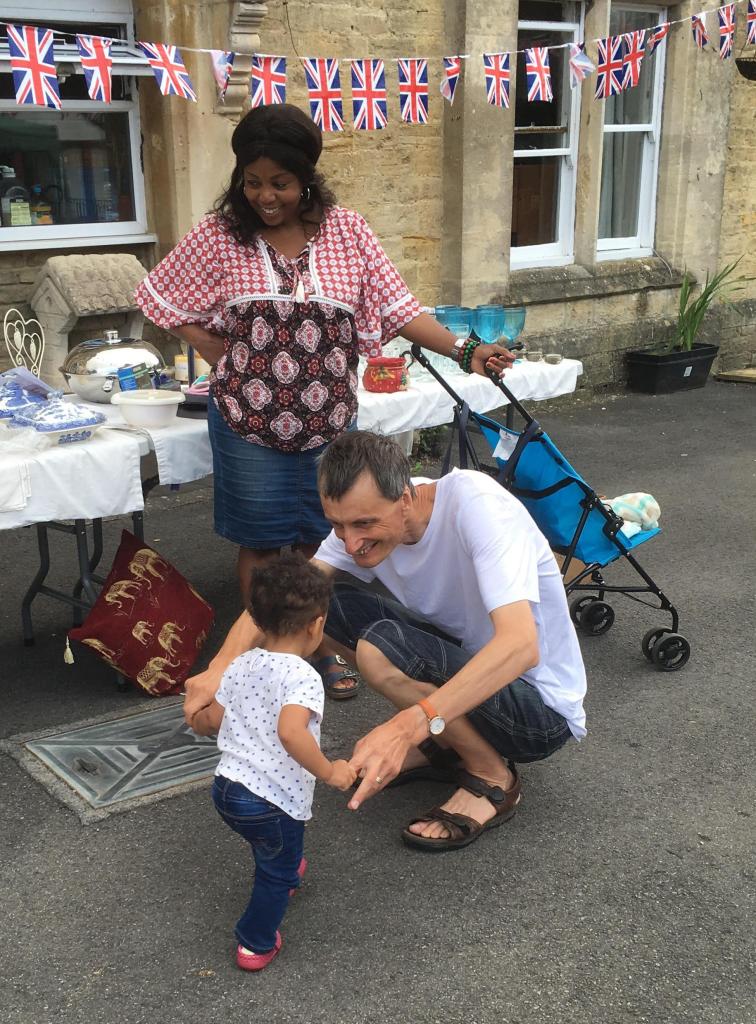 a child walking up to a man that is crouched down waiting for the child with a lady stood up looking at them