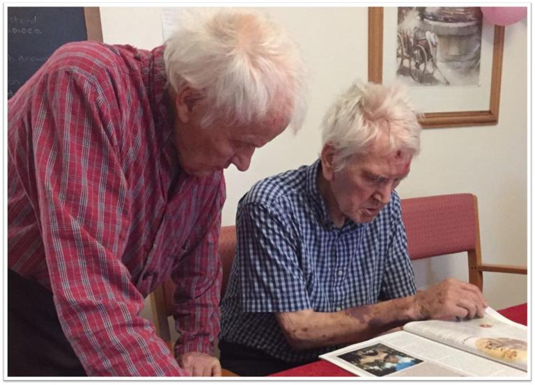 two elderly men on sat one standing reading a magazine