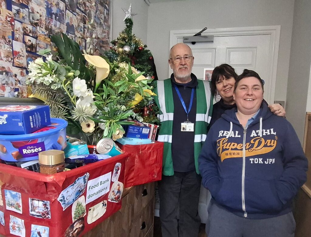 Three adults standing beside a Christmas donation collection at Frome Nursing Home, with a decorated tree behind them. A table in front holds food bank donations including chocolates, tins, toiletries and festive items, with a sign reading “Food Bank donations”.